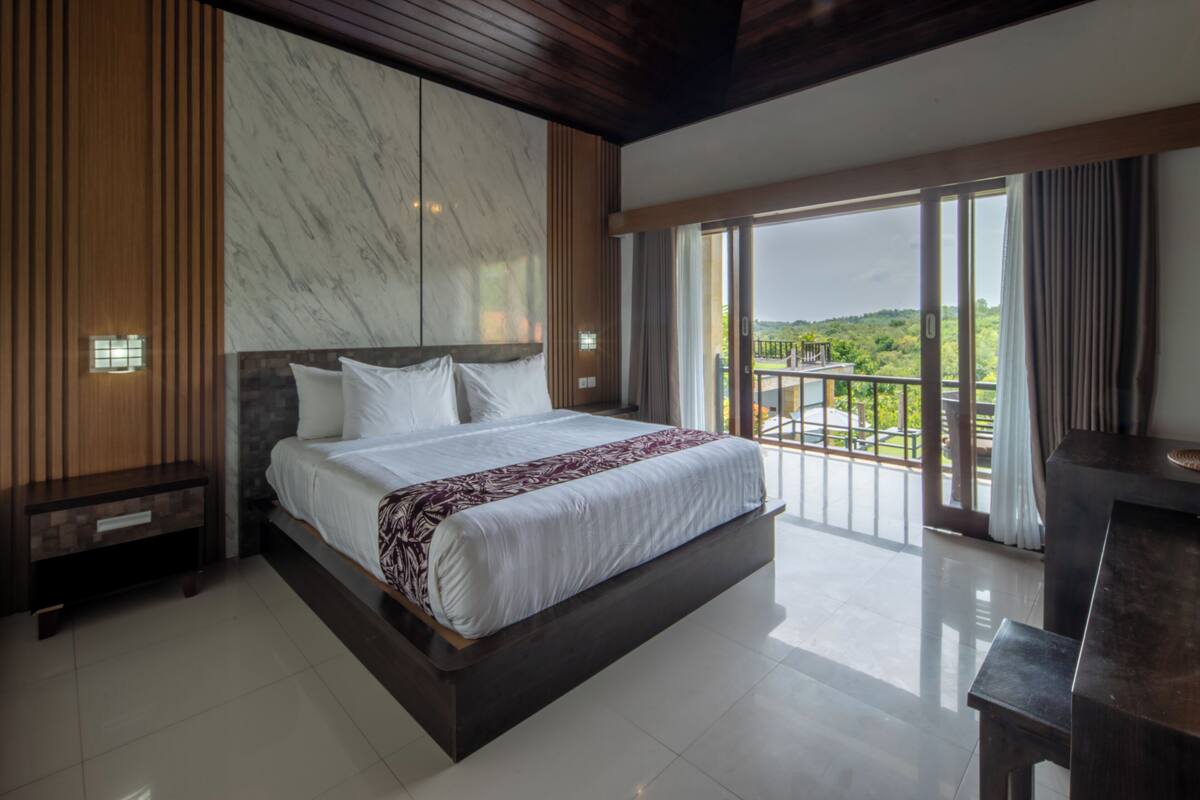 A spacious bedroom with a large bed, wooden paneling, a marble accent wall, and a balcony with a view.