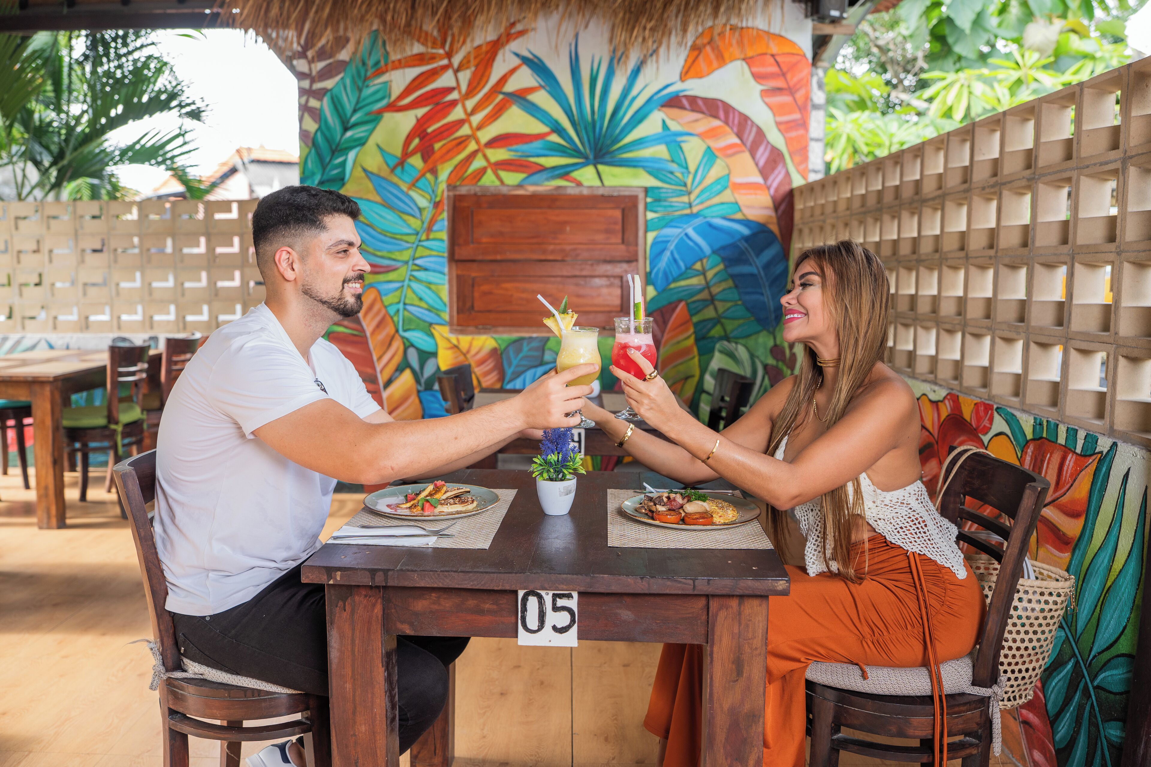 A wooden table with a number 05, two people seated, holding drinks, and a colorful mural in the background.