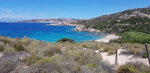 A coastal landscape with a sandy beach, rocky outcrops, and sparse vegetation.
