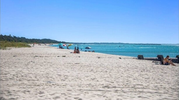 Una playa con gente relajándose, sombrillas y un muelle al fondo.