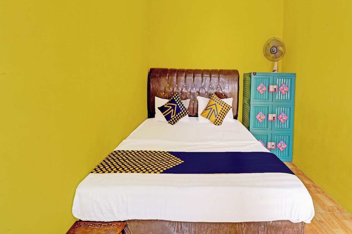 Una cama con cabecera de madera, varios almohadones, una colcha azul y blanca, y un aparador azul verdoso con adornos rosados.