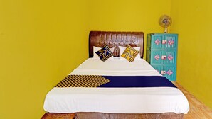 Een bed met een wit- en blauwe spreideken, twee kussens met geometrische patronen, een houten hoofdbord en een zeegroene kast met roze en gele decoraties.