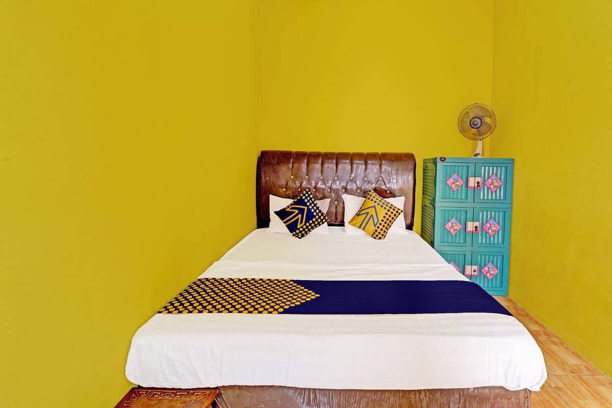 A bed with a white and blue bedspread, two pillows with geometric patterns, a wooden headboard, and a teal cabinet with pink and yellow designs.