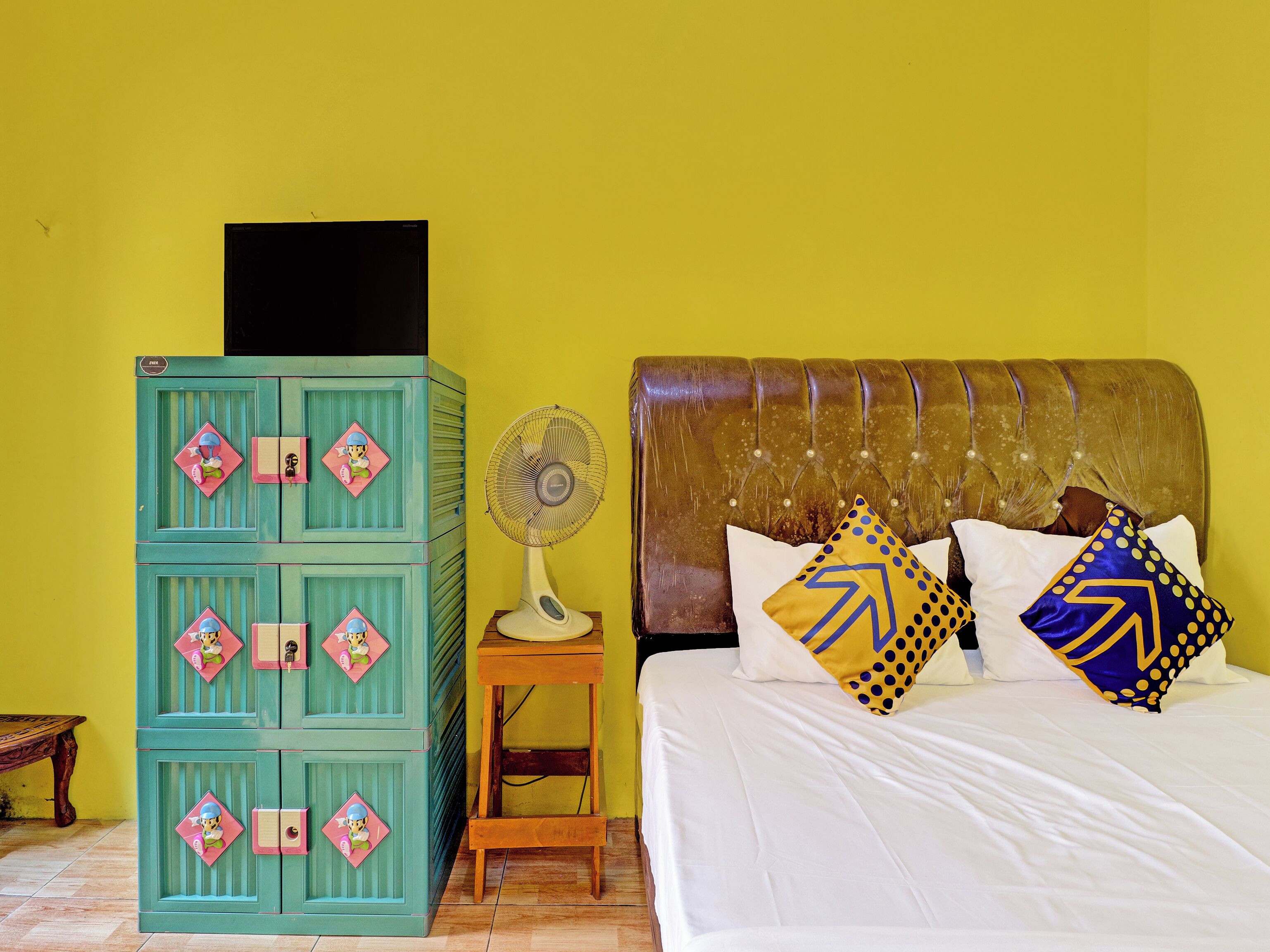 A room with a bed, a chest of drawers, a fan, and a small table.