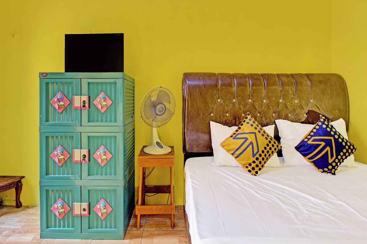 A room with a bed, a chest of drawers, a fan, and a small table.