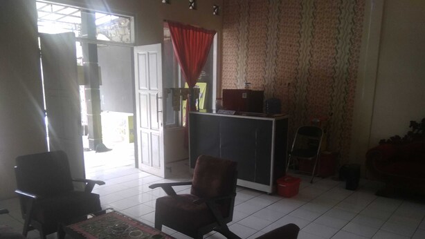 Una habitación con mostrador de recepción, dos sillas, una mesa con mantel estampado y suelo de baldosas.