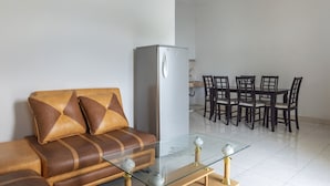 Een woonkamer met een bank, een glazen salontafel, een koelkast, en een eethoek met stoelen en een tafel.