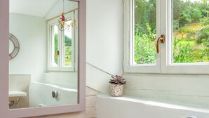 Une salle de bain avec une baignoire blanche, un miroir et une fenêtre donnant sur un paysage verdoyant.