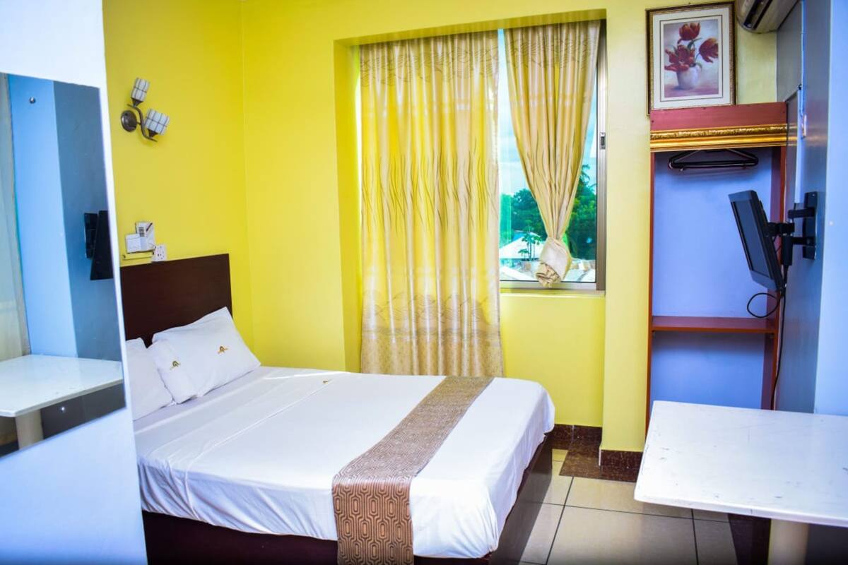 Une chambre d’hôtel avec un lit, un bureau, une fenêtre avec des rideaux, un téléviseur fixé au mur et une petite étagère sur laquelle se trouve un cadre photo.