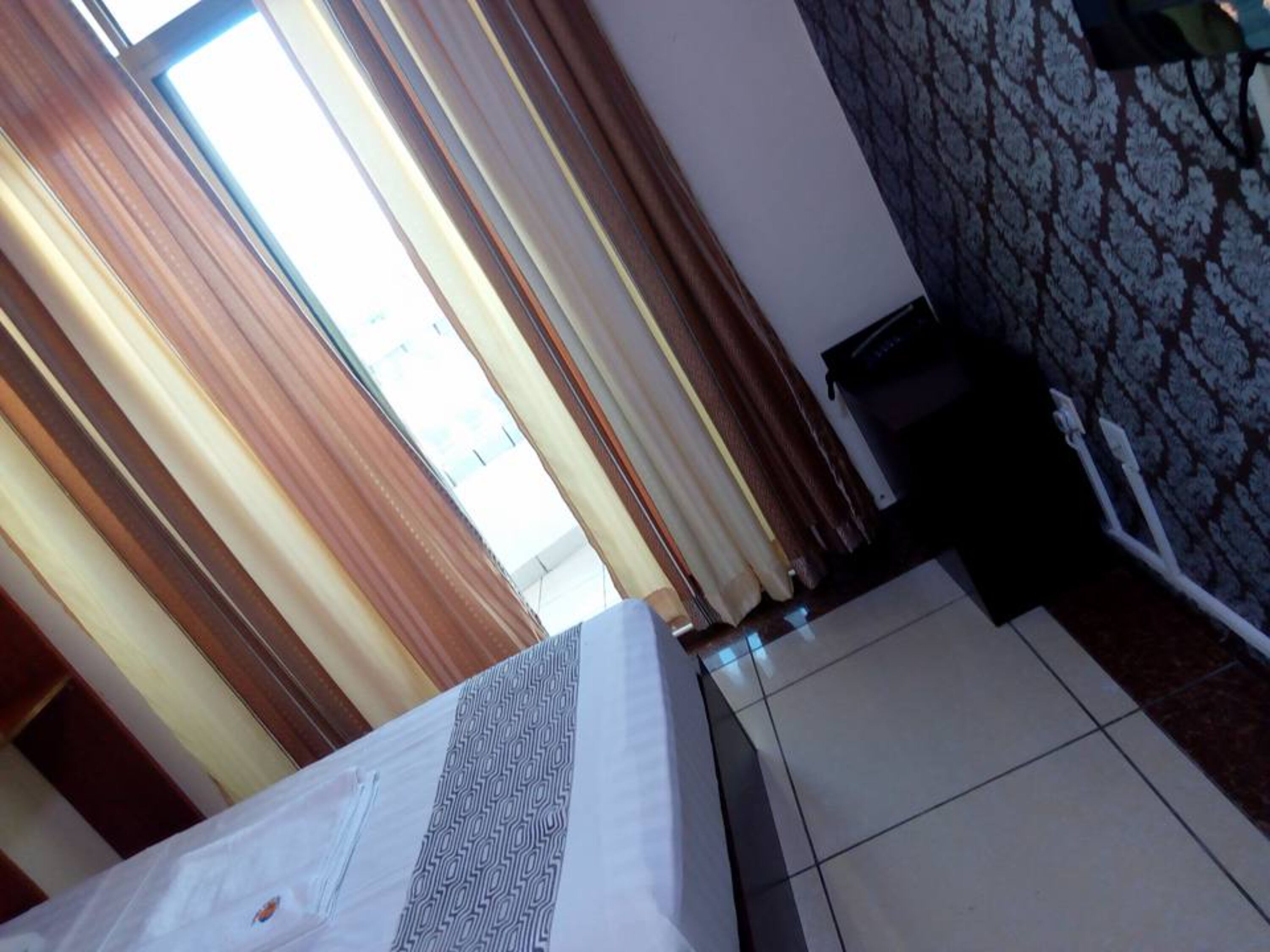 A hotel room with a bed, curtains, a tiled floor, and a patterned wall.