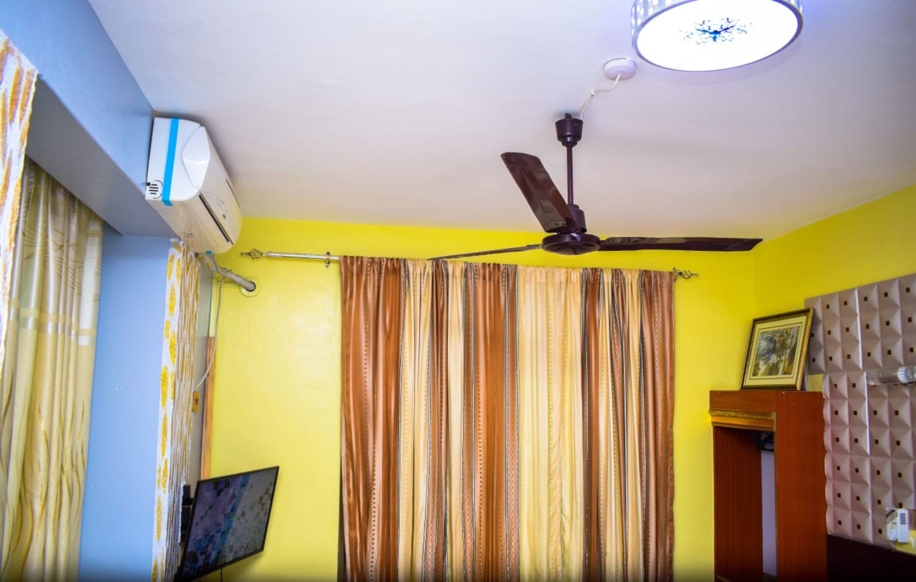 Una habitación con ventilador de techo, equipo de aire acondicionado, cortinas a rayas y un cuadro sobre un atril.