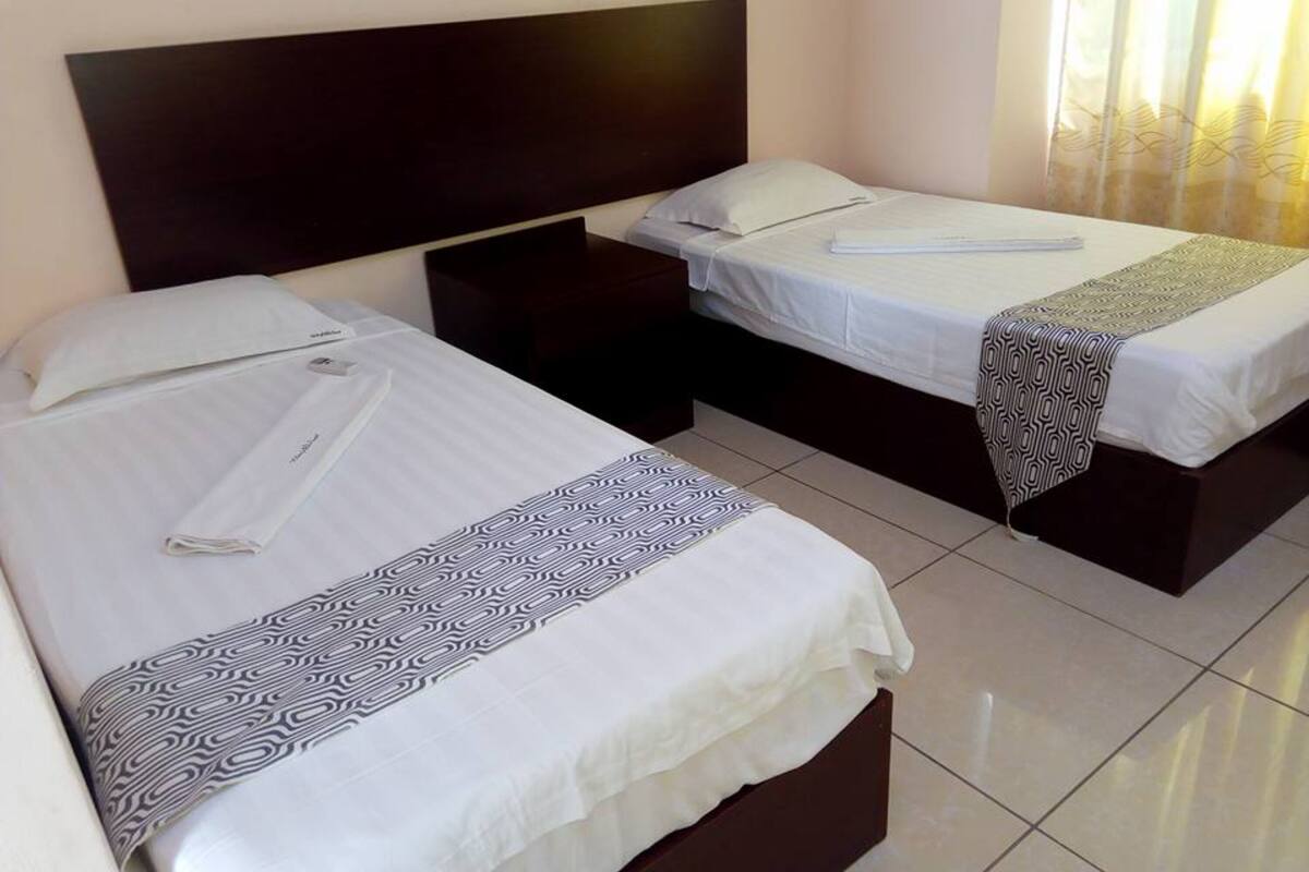 Une chambre d’hôtel avec deux lits, une tête de lit sombre, une table de chevet et une fenêtre avec des rideaux.