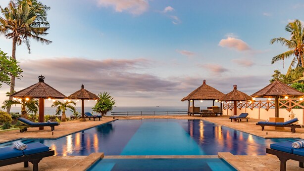 A pool area with lounge chairs and thatched-roof gazebos, overlooking a body of water.