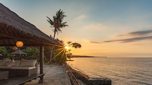 A seaside patio with a thatched roof, wicker furniture, and a view of the sunset over the ocean.