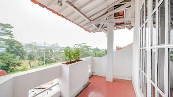 A balcony with a white railing, a red-tiled floor, and a view of greenery and buildings in the distance.