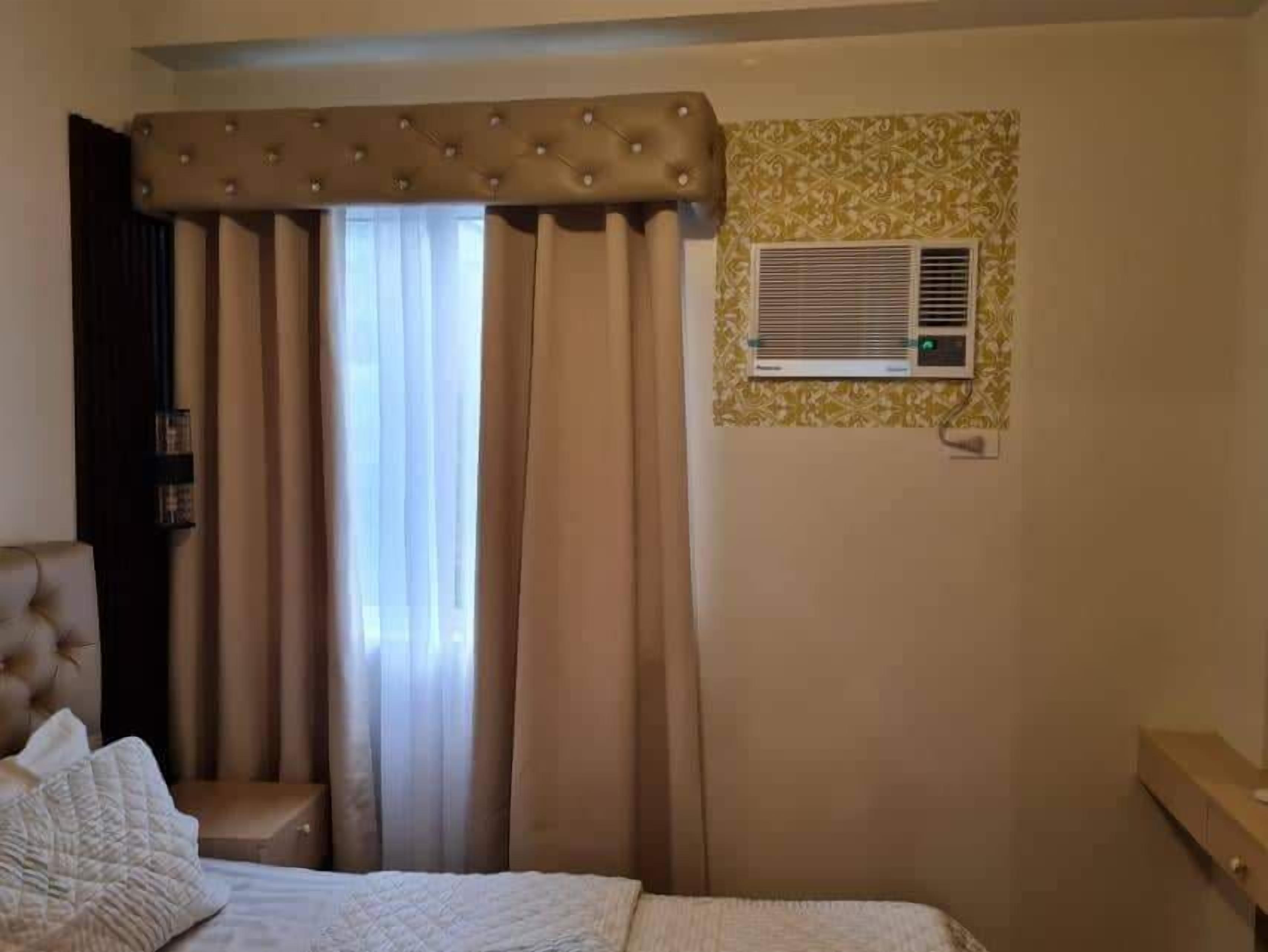 A hotel room with a bed, a headboard, a window with curtains, and a wall-mounted air conditioning unit.