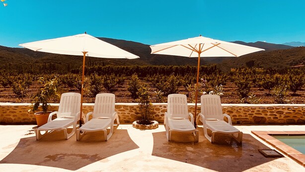 Area piscina con poltronne da relax bianche e ombrelloni, un muro in pietra e vista sui vigneti e le montagne sullo sfondo.