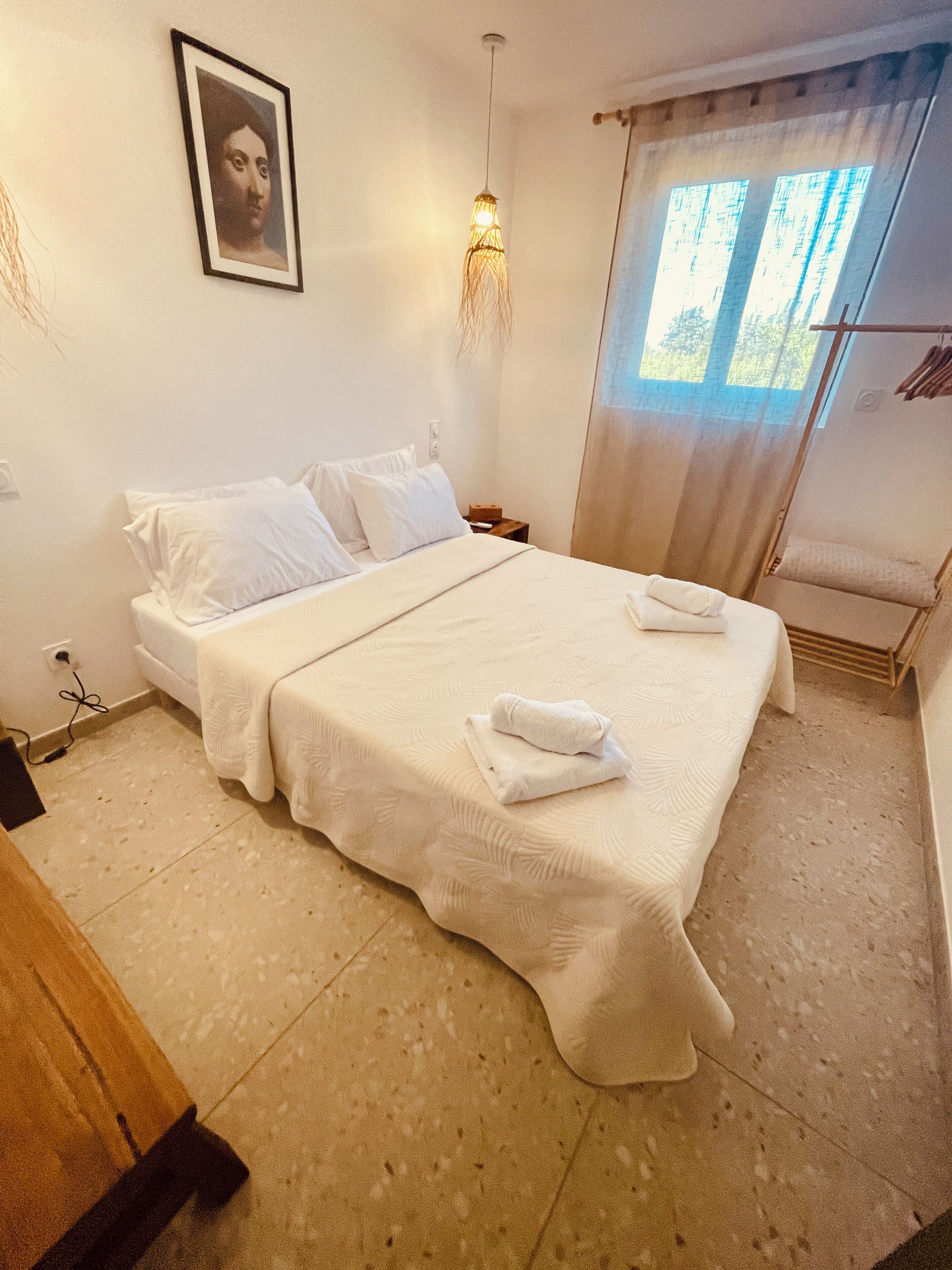 A neatly made bed with white linens and a folded towel, a bedside table with a lamp, and a window with sheer curtains.