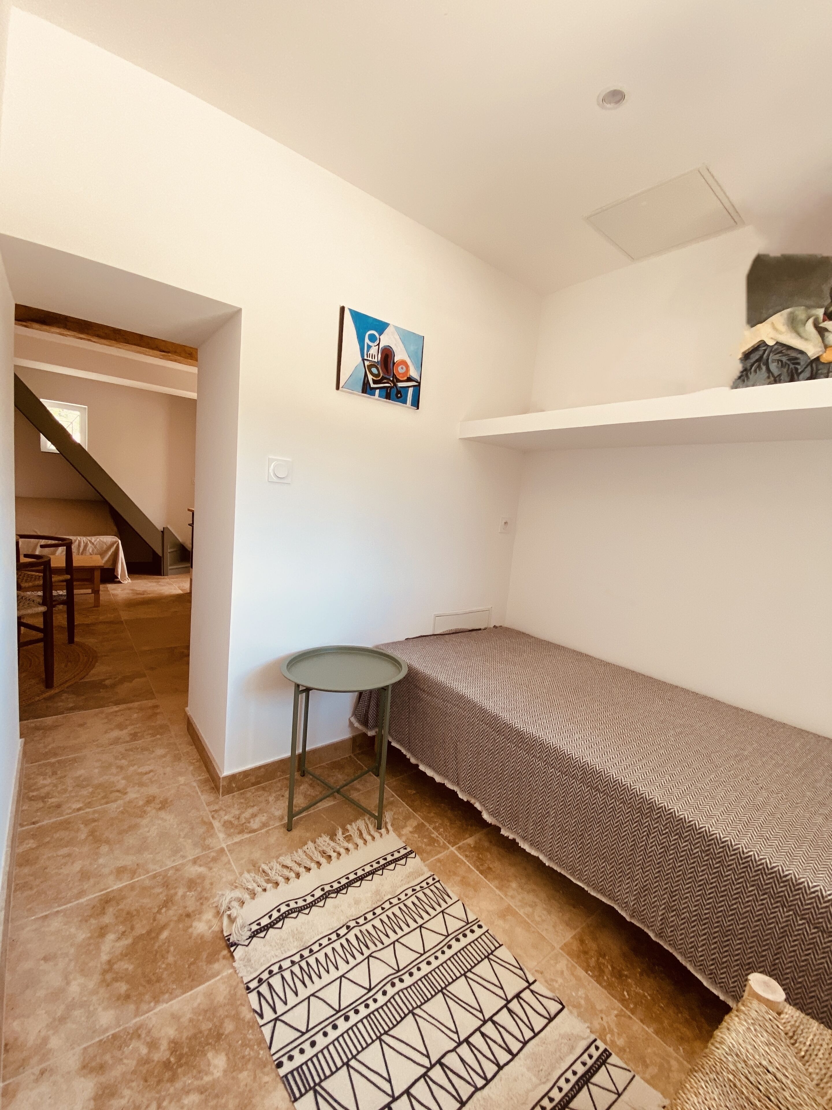A small, sparsely furnished room with a single bed, a small table, and a rug.