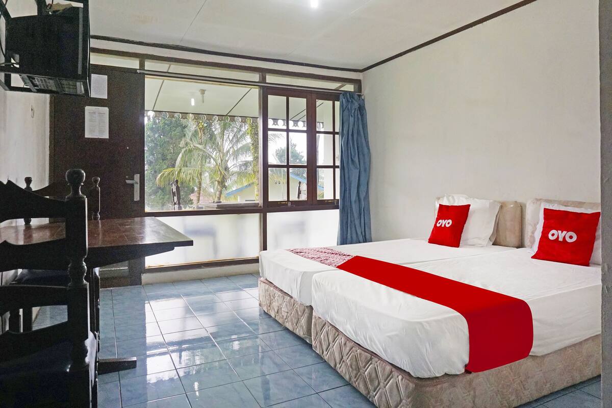 Una habitación de hotel con cama, escritorio, televisor y ventana con vista a palmeras.