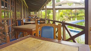 Un área de comedor al aire libre techada con mesas y sillas de madera, vista a una piscina y un edificio al fondo.