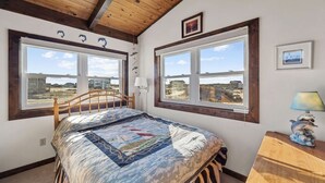 A bedroom with a wooden ceiling, large windows with views of the beach, a bed with a patterned bedspread, and a small table with a lamp.
