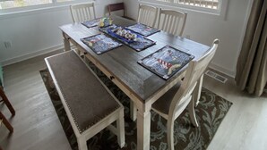 A dining area with a wooden table, white chairs, and a ceiling fan. There are windows with views of the beach and a framed quote on the wall.