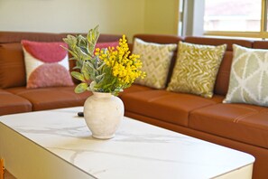 Un jarrón blanco con flores amarillas sobre una mesa blanca en un salón con un sofá marrón y cojines estampados.