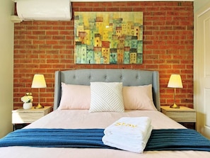 Una habitación de hotel con pared de ladrillo, una cama con cabecera gris, dos mesitas de noche con lámparas y un cuadro sobre la cama.