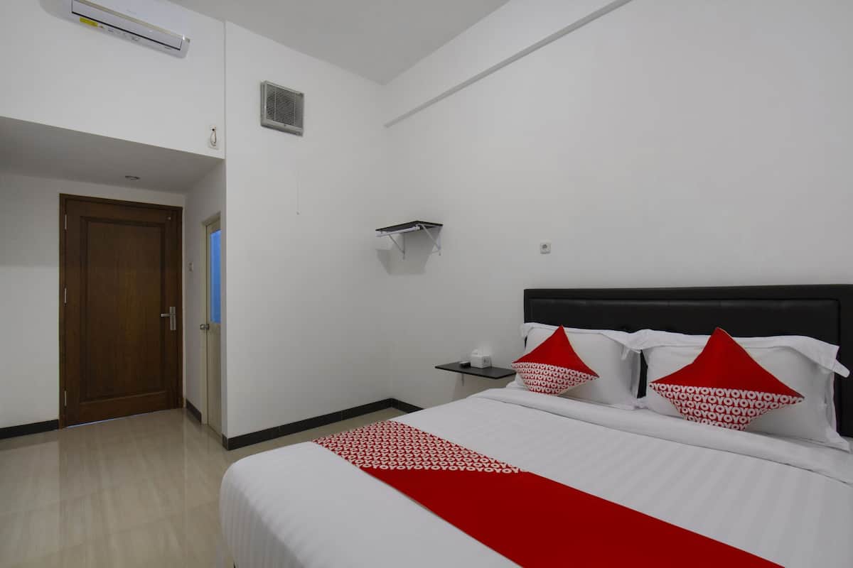 Quarto de hotel com uma cama, uma porta de madeira, uma mesinha e uma prateleira fixada na parede.