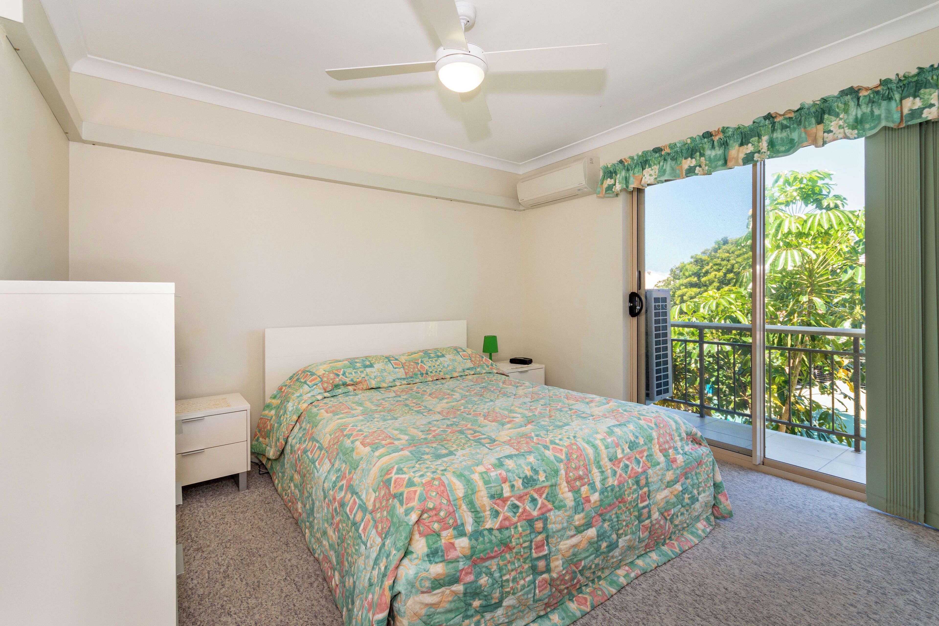 A bedroom with a bed, a nightstand, a ceiling fan, and a sliding glass door leading to a balcony with a view of greenery.