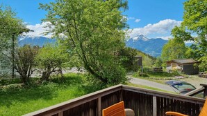 Eine Terrasse mit Holzstühlen und einem Tisch, mit Blick auf eine Berglandschaft mit See und einer Hütte.