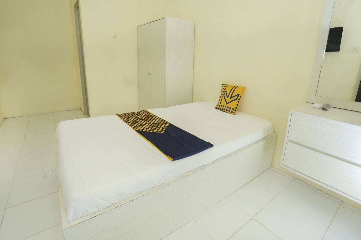 Une chambre avec un lit simple, recouverte d’une housse de couette blanche, d’une couverture bleue et jaune, et comprenant une armoire blanche et un miroir au mur.