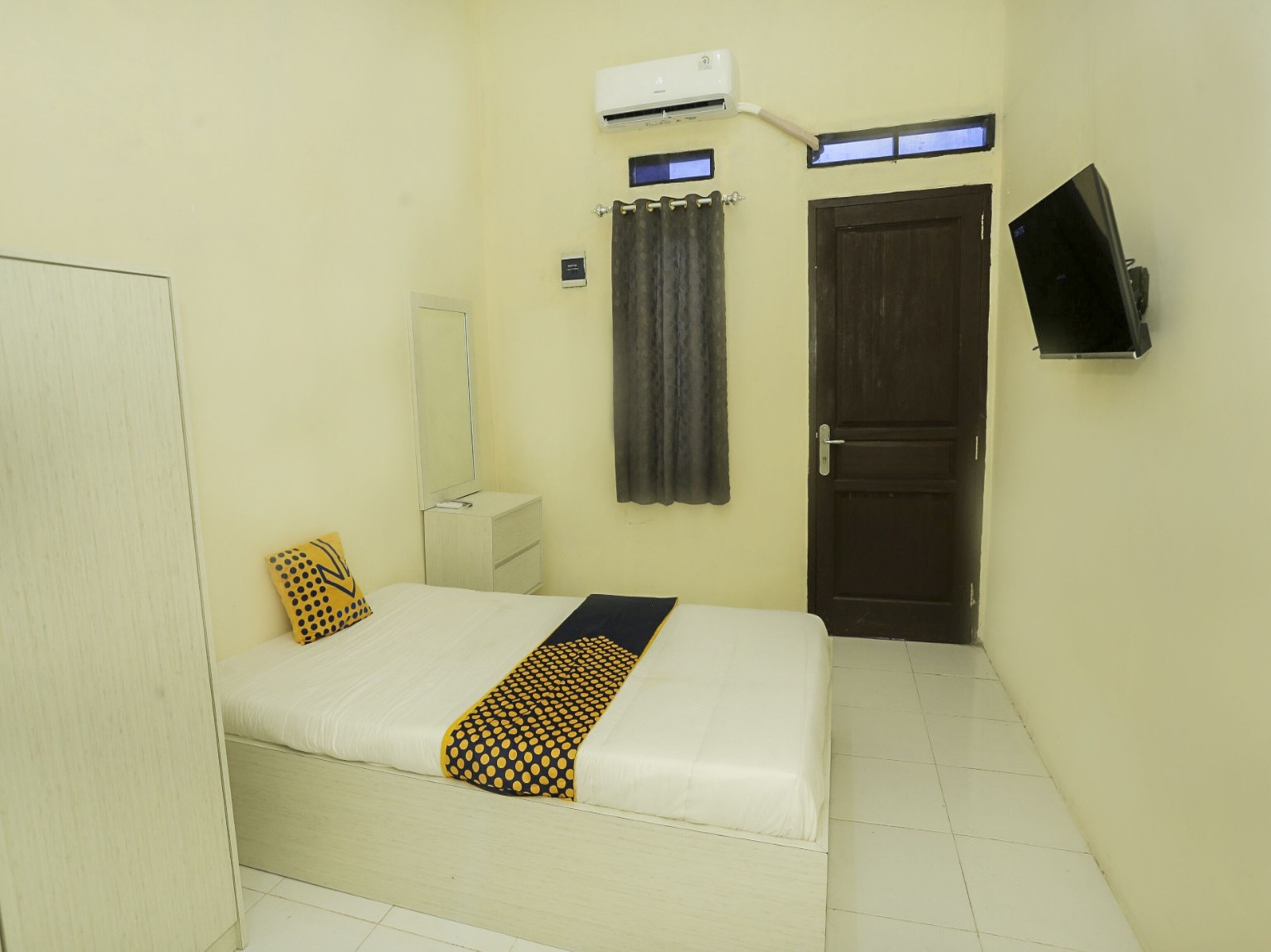 A small, clean room with a bed, a bedside table, a mirror, an air conditioning unit, a door, and a TV mounted on the wall.
