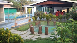 A modern outdoor pool area with a covered seating area, surrounded by potted plants and trees. - Canal Touch Resort (Karjat)