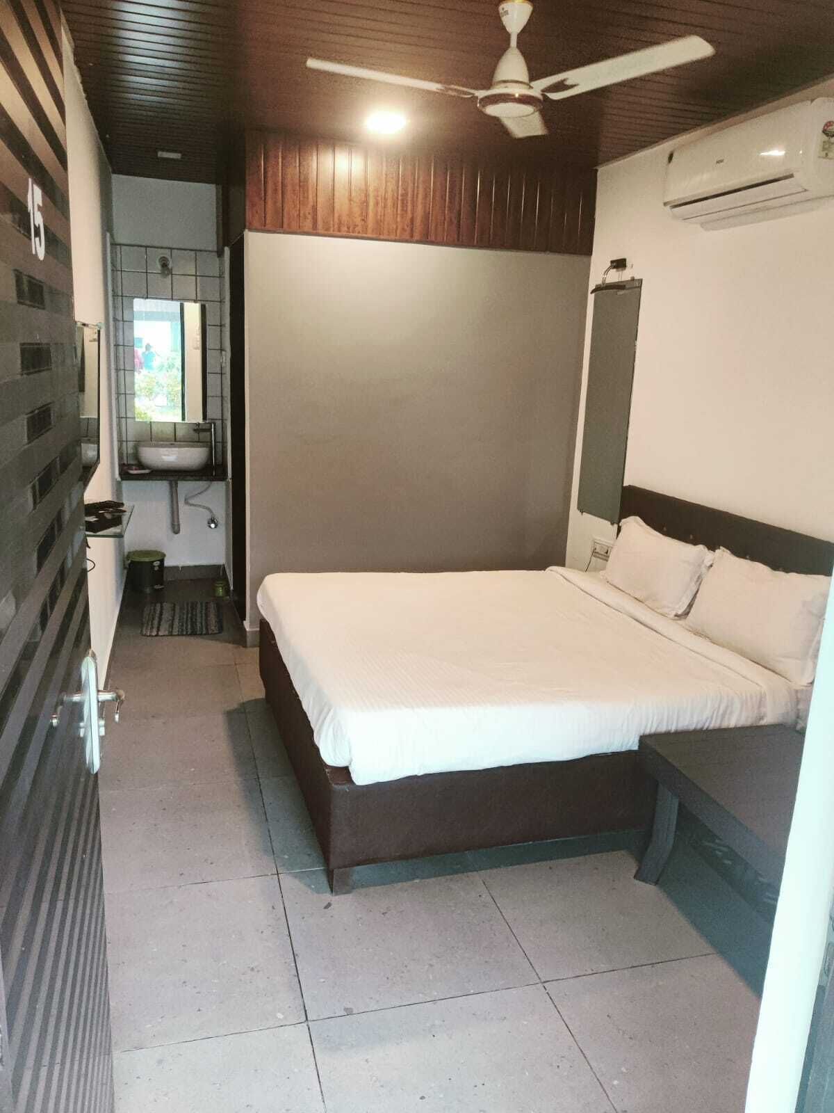 A bedroom with a large bed, a ceiling fan, an air conditioning unit, and a small bathroom with a sink and mirror.