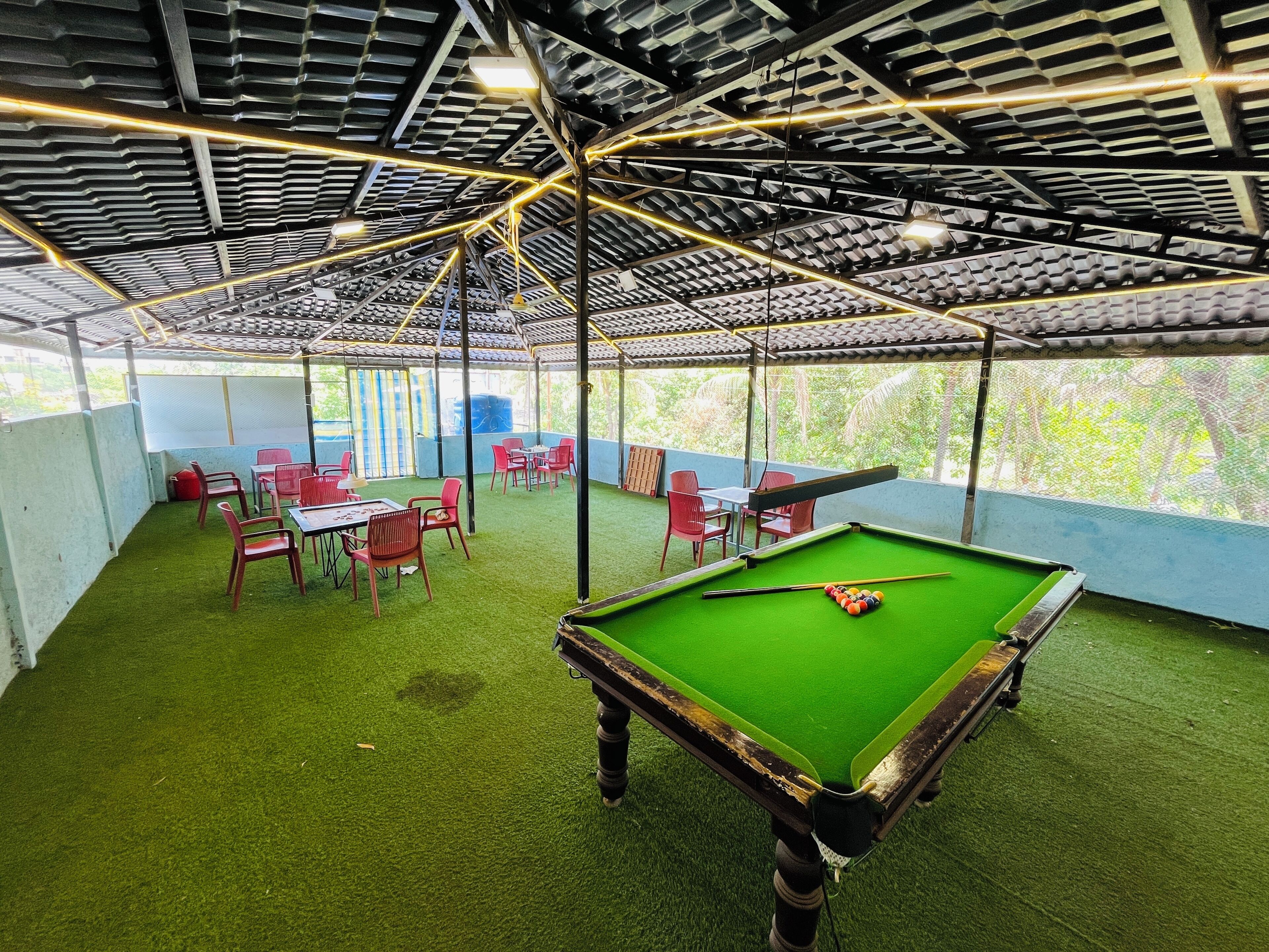 A pool table with a green felt surface, surrounded by red chairs and tables, under a metal roof structure.