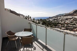 A balcony with a round table and chairs, offering a view of a coastal town and the sea. - Servatur Castillo de Sol (Mogan)