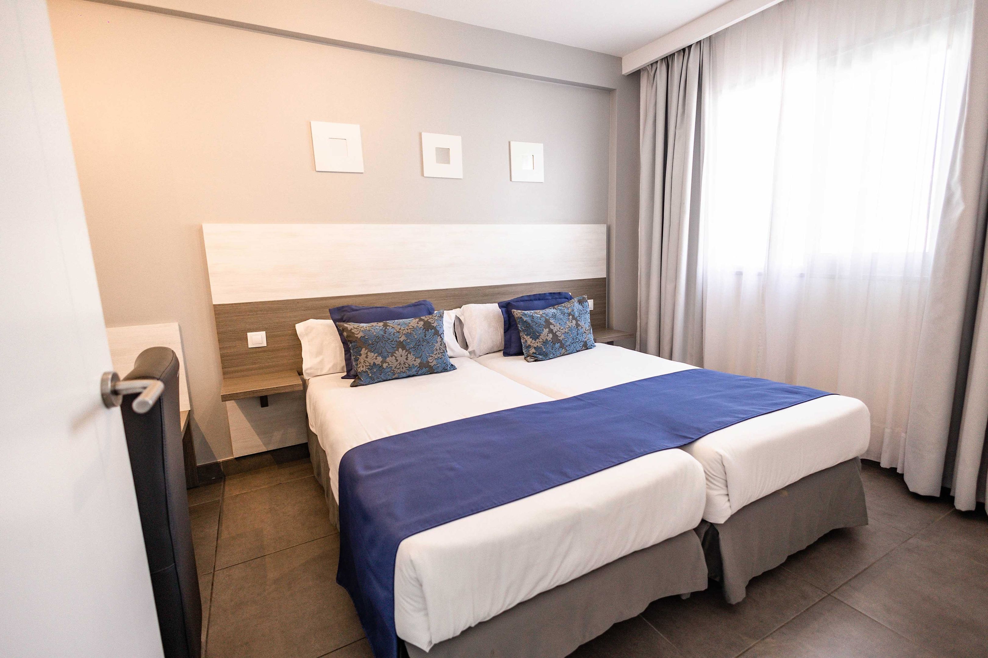 Una cama doble con colcha azul y blanca, cabecera de madera y ventana con cortinas.
