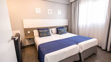 Una cama doble con colcha azul y blanca, cabecera de madera y ventana con cortinas.