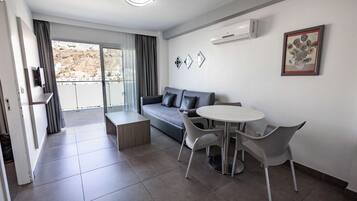 A modern living room with a grey sofa, white round table, and matching chairs. There is a balcony with a view of buildings and a mountain.