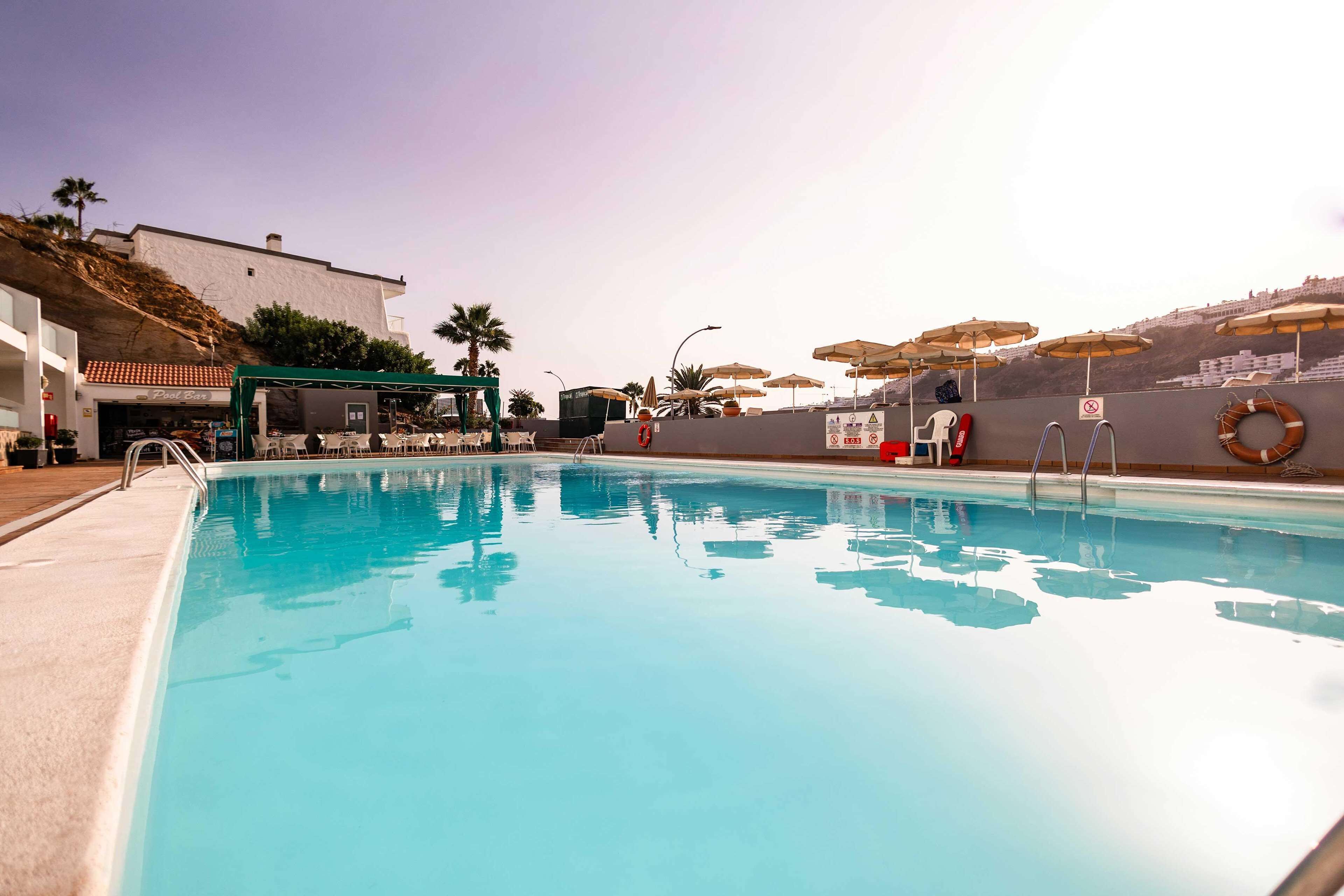 A clear swimming pool with a lifeguard chair and umbrellas, a building with a red-tiled roof, and palm trees in the background.