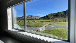 A room with a large window offering a view of a green field, a mountain, and a clear blue sky. - Pudding Hill Lodge & Chalets (Methven)