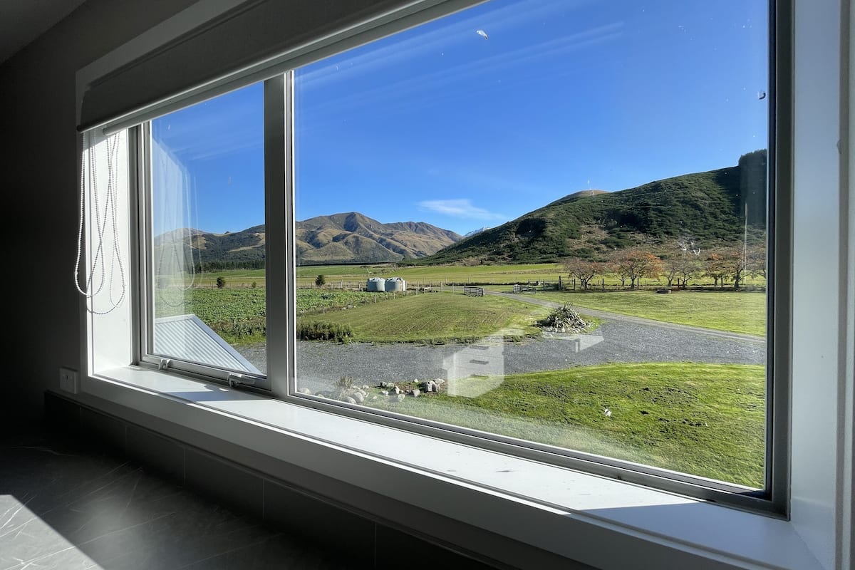 A room with a large window offering a view of a green field, a mountain, and a clear blue sky.