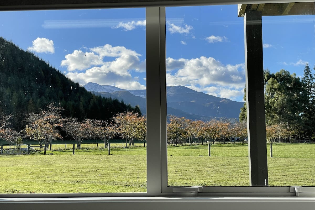 A view through large windows of a grassy field with trees and mountains in the background.