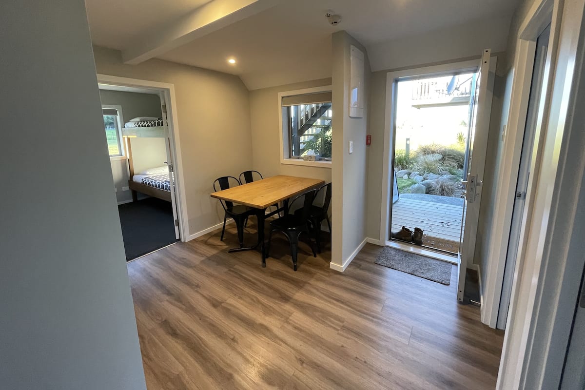 A room with a wooden floor, a dining table with four chairs, a bunk bed, and a sliding glass door leading to an outdoor area.