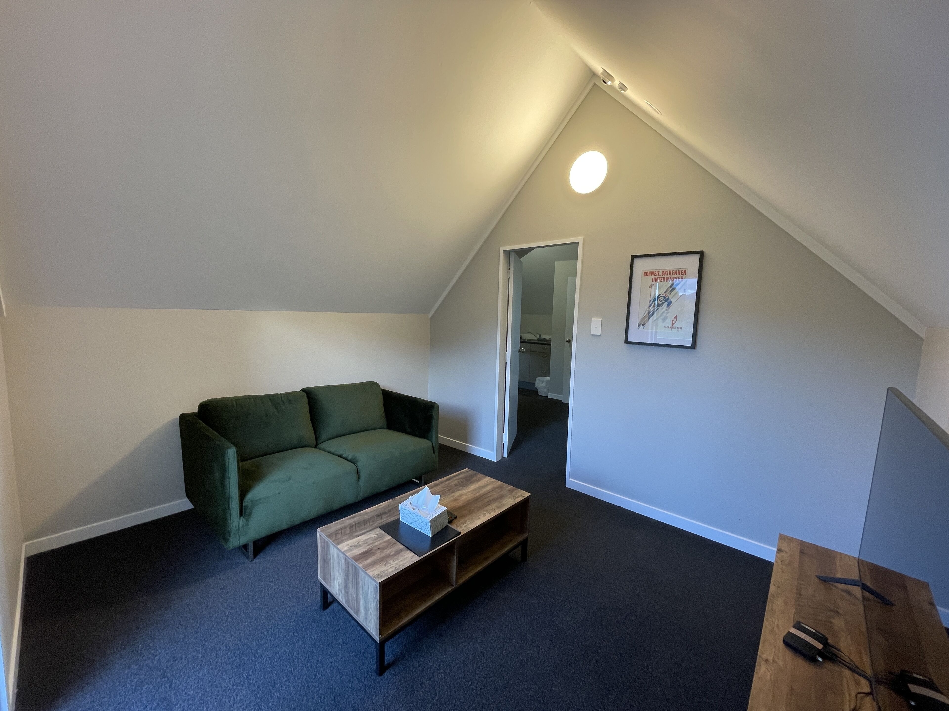 Een kamer met een laag schuin plafond, een groene bank, een houten bijzettafeltje en een ingelijst kunstwerk aan de muur.