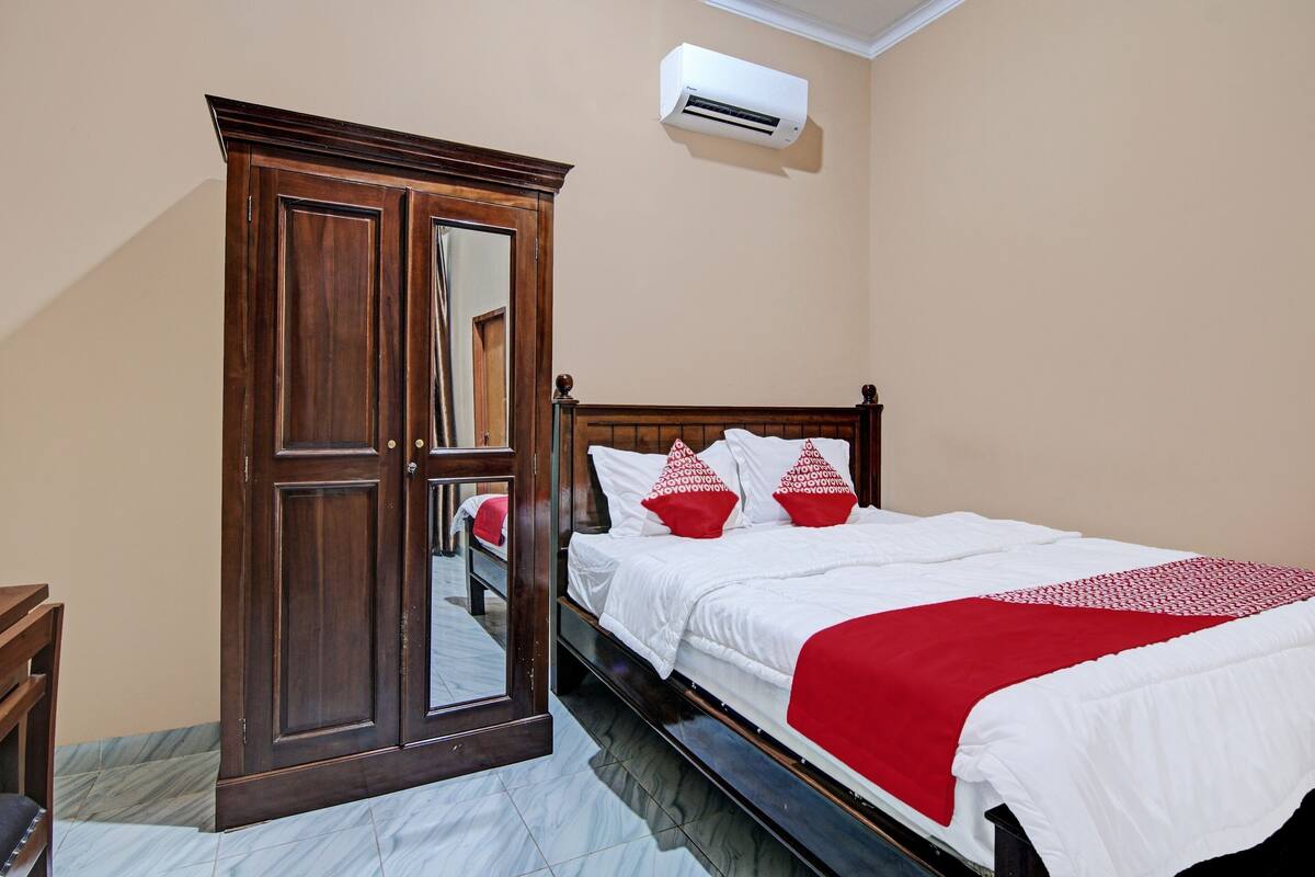 A bedroom with a wooden wardrobe, a bed with white bedding and red accents, and an air conditioning unit on the wall.