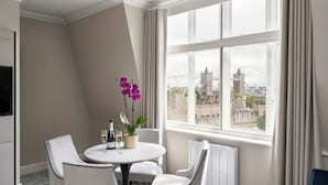 Ett modernt hotellrum med ett runt matbord, vita stolar, en orkidé i en kruka och utsikt över ett historiskt torn genom stora fönster.