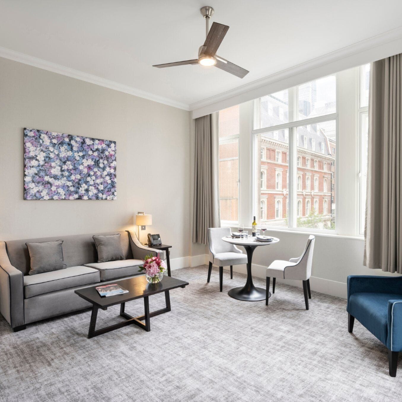 Ein modernes Wohnzimmer mit einer Couch, einem Couchtisch, einem Esstisch mit Stühlen, einem Deckenventilator und einem großen Fenster mit Blick auf die Stadt.